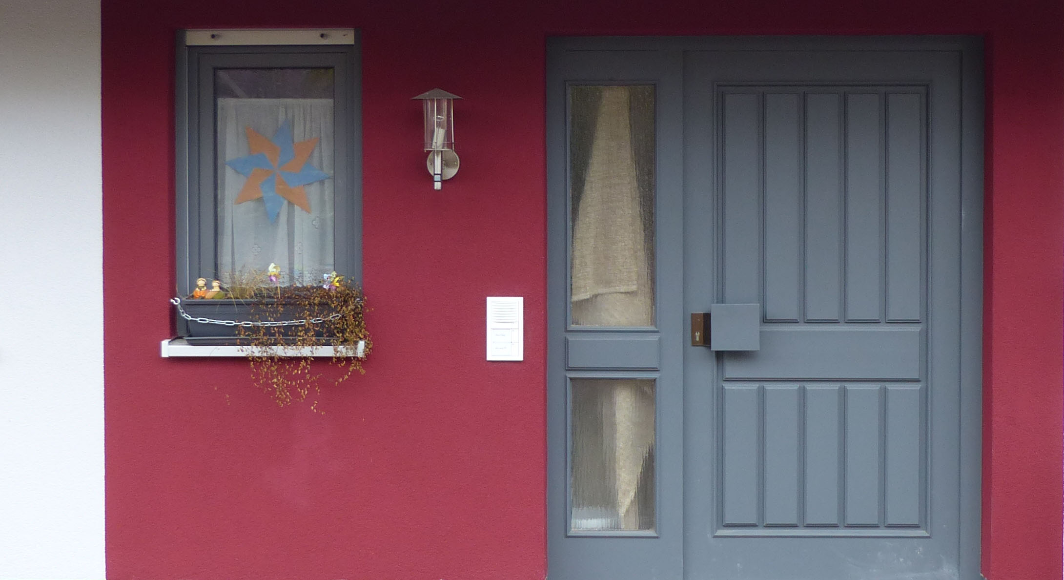 Eingangsbereich mit grauer Haustür mit Seitenelement, Fenster mit grauem Rahmen, weinrot verputzte Wand.
